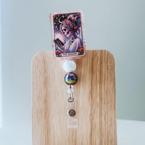 May include: A retractable badge reel with a tarot card design. The card features a skeleton figure reading a book, with the text "THE READER". The reel has a white bead, a color-shifting bead, and a clear plastic clip.
