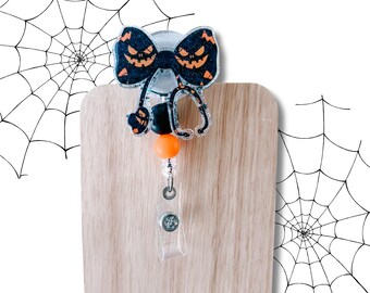 Puede incluir: Un portatarjetas con temática de Halloween con un lazo negro con caras de calabaza naranja. El carrete incluye un diseño de estetoscopio, una cuenta naranja y un clip de plástico transparente. El fondo tiene un diseño de telaraña.