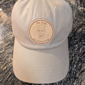 May include: A beige baseball cap with a brown leather patch that says "John Brown Did Nothing Wrong" and features a black and white illustration of a man.