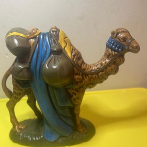 May include: A ceramic camel figurine, painted in shades of brown, tan, and gold, with a blue blanket and saddlebags. The camel has a blue headpiece and is standing on a green base. A decorative item.