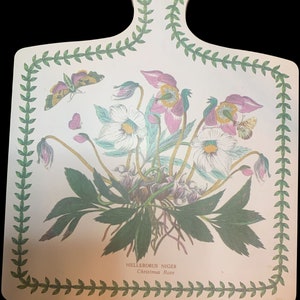 May include: A white wooden cutting board with a green border and a floral design featuring white and pink flowers, green leaves, and butterflies. The text "Helleborus Niger Christmas Rose" is printed on the board.