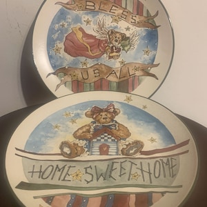 May include: Two decorative ceramic plates with a patriotic theme. The top plate features a teddy bear angel with wings and a banner that reads "Bless Us All". The bottom plate features a teddy bear sitting on a house with a banner that reads "Home Sweet Home". Both plates have a blue, red, and white striped background with stars.