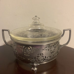 May include: Silver serving dish with a glass lid. The dish has a cut-out design of flowers and leaves. The dish sits on three feet.