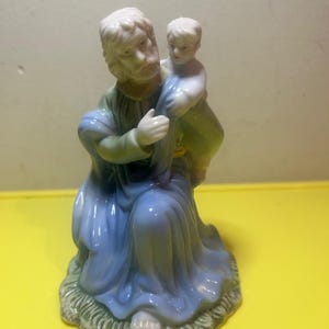 May include: A ceramic figurine depicting a seated figure in blue and green robes holding a child. The figurine has a detailed face and is set on a base with a grass-like texture. The color palette includes shades of blue, green, and white.