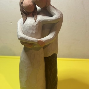 May include: A wooden figurine depicting a couple embracing. The woman wears a light-colored dress, while the man has dark pants and a light-colored shirt. The carving style gives a rustic, handmade feel.