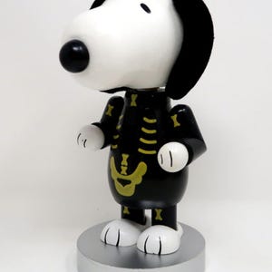 May include: A Snoopy figurine featuring a white head with black ears and a black nose, wearing a black outfit with a yellow skeleton bone print. The character stands on a round silver base, with white paws and a smooth, glossy finish throughout.