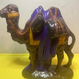 May include: A brown ceramic camel figurine with a purple saddle and two black bags. The camel has yellow and brown accents on its harness and saddle. The figurine is standing on a green base.