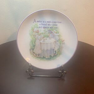 May include: A white ceramic plate with a purple border features a painting of two girls having tea in a garden. The plate has the text "A sister is a wish-come-true... a friend who cares and shares with you!"