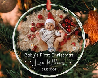 May include: A round, clear acrylic Christmas ornament featuring a photo of a baby in a Santa hat and polka dot onesie surrounded by red ornaments. The text reads Baby's First Christmas, Liam Williams, and the date 08.15.2026.