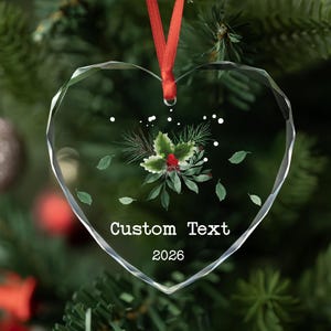 May include: A clear, heart-shaped glass ornament with faceted edges, suspended by a red ribbon. The center features a printed illustration of green holly leaves, pine needles, and red berries, with the text Custom Text and 2026 below.