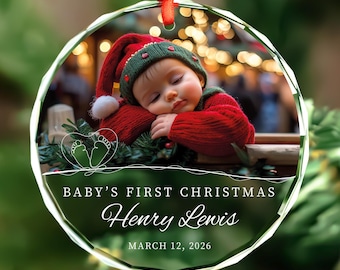May include: A round, faceted glass Christmas ornament featuring a photo of a baby in a red and green hat sleeping on pine branches. The text reads BABY'S FIRST CHRISTMAS, Henry Lewis, and MARCH 12, 2026, with a white line drawing of baby feet.