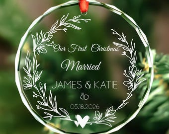 以下が含まれることがあります： 赤いリボンが付いた、透明な丸いクリスマスオーナメント。「Our First Christmas Married」の文字と「JAMES & KATIE」の名前、結婚式の日付05.18.2026が、花のリースデザインで囲まれています。