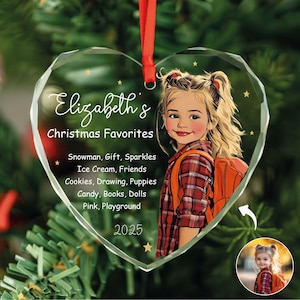 May include: Heart-shaped Christmas ornament with a portrait of a girl wearing a plaid shirt and an orange backpack. The ornament has the text "Elizabeth's Christmas Favorites" and a list of favorite things. A red ribbon is attached for hanging.