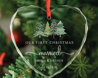 May include: Heart-shaped clear glass Christmas ornament with a red ribbon. The ornament features a watercolor design of evergreen trees and the text "OUR FIRST CHRISTMAS married" with names and a date.