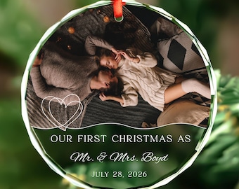 May include: A round, faceted glass Christmas ornament featuring a photograph of a couple lying together. The ornament includes a white line-art heart and the printed text: OUR FIRST CHRISTMAS AS Mr. & Mrs. Boyd JULY 28, 2026, with a red ribbon.