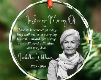 Puede incluir: Un adorno conmemorativo circular de cristal con borde facetado, colgado de una cinta roja. Presenta un retrato en blanco y negro de una persona con bufanda, junto al texto In Loving Memory Of, un poema, el nombre Isabella William y 1965-2026.