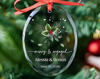May include: An oval-shaped clear glass ornament with faceted edges, featuring a red ribbon hanger. The center displays a watercolor holly sprig, white text reading "merry & engaged", "Alessia & Ronan", and the date "June 10, 2026".