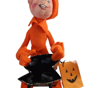 May include: An orange felt doll with a smiling face, wearing a matching hood and outfit. The doll holds a black and silver IKEA shopping bag and a small orange paper bag with a jack-o'-lantern face.