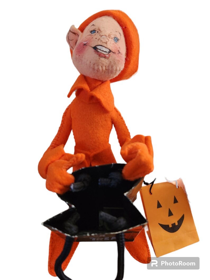 May include: An orange felt doll with a smiling face, wearing a matching hood and outfit. The doll holds a black and silver IKEA shopping bag and a small orange paper bag with a jack-o'-lantern face.
