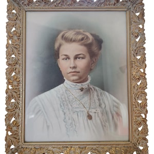 May include: Portrait of a woman in a white dress with lace detailing, framed in an ornate gold-colored frame. The portrait features a woman with light skin, blonde hair, and a gold pendant necklace. The frame has a detailed floral and leaf design.