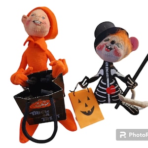 May include: Two Halloween dolls. One is dressed in orange with a matching hat and holding a black bag with the words "Trick or Treat" and a jack-o'-lantern. The other doll is dressed as a skeleton with a black top hat, holding a walking stick and a jack-o'-lantern bag.