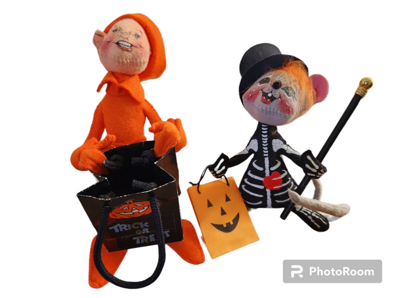 May include: Two Halloween dolls. One is dressed in orange with a matching hat and holding a black bag with the words "Trick or Treat" and a jack-o'-lantern. The other doll is dressed as a skeleton with a black top hat, holding a walking stick and a jack-o'-lantern bag.