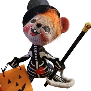 May include: A Halloween felt mouse doll in a black top hat and skeleton costume. The mouse holds a black cane with a gold top and an orange jack-o'-lantern bag. The mouse has orange hair and a painted face with red cheeks.