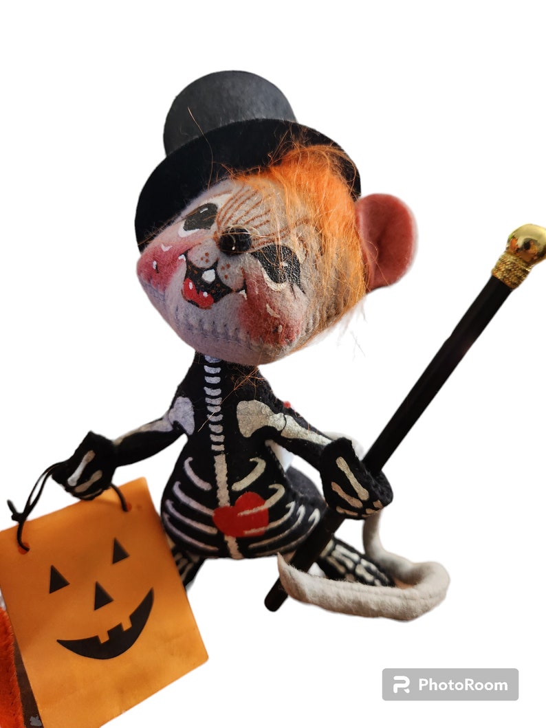 May include: A Halloween felt mouse doll in a black top hat and skeleton costume. The mouse holds a black cane with a gold top and an orange jack-o'-lantern bag. The mouse has orange hair and a painted face with red cheeks.