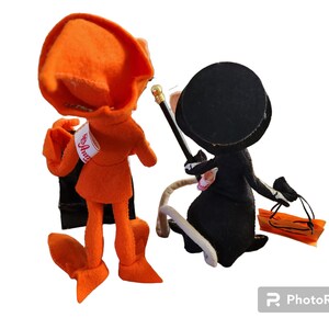 May include: Two handmade felt dolls. One doll is orange with a matching hat and black cape. The other doll is black with a top hat, holding a cane and a small orange bag. The dolls are standing next to each other.