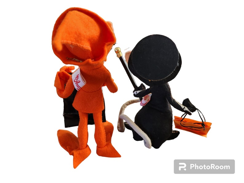 May include: Two handmade felt dolls. One doll is orange with a matching hat and black cape. The other doll is black with a top hat, holding a cane and a small orange bag. The dolls are standing next to each other.