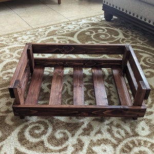 May include: A dark brown wooden dog bed with a slatted bottom. The bed is empty and sitting on a patterned rug.