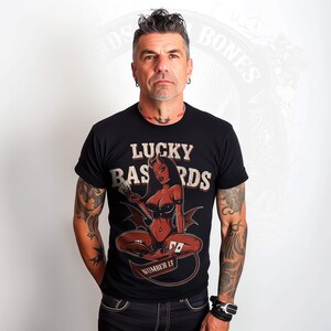 Pode incluir: Uma camiseta preta com uma estampa gráfica de uma mulher com chifres de diabo e cauda, segurando um cartão de jogo. O texto "LUCKY BASTARDS" está acima da mulher e "NUMBER 13" está abaixo dela.