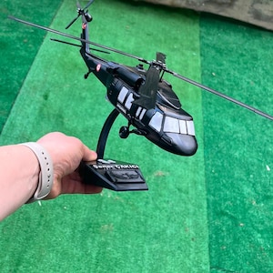 May include: A black scale model helicopter featuring a main rotor and tail rotor, mounted on a black display stand. The fuselage displays the text JANDARMA and the base features a plaque with the name Sener ÇAKICI and an emblem.