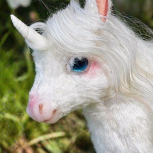 May include: A detailed image of a white unicorn with a spiralled horn, blue eyes, and pink details on its ears and snout. The unicorn has long, flowing white mane and is set against a blurred green and white backdrop.