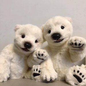 May include: Two white plush polar bear stuffed animals sitting side by side. The bear on the left is looking to the right and the bear on the right is looking to the left. Both bears have black eyes and noses and black paw pads.