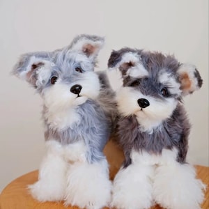 Op de afbeelding: Twee pluche Schnauzer hondenknuffels. De honden hebben grijs en wit haar, zwarte neuzen en bruine ogen. Ze zitten op een houten oppervlak. De knuffels zijn gedetailleerd en realistisch.