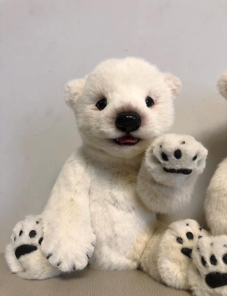May include: A white plush polar bear teddy with black eyes, nose, and paw pads. The bear is sitting with its right paw raised in a waving gesture.
