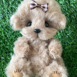 May include: A plush, light brown teddy bear with a small, brown and white checkered bow on its head. The bear has a black nose, dark eyes, and detailed paws. The fur is soft and fluffy, giving it a cuddly appearance.
