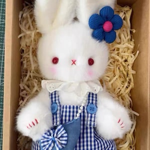 May include: A plush white bunny doll with a blue flower hair accessory and red eyes. The bunny wears blue and white checkered overalls with a matching bow and a small blue purse. The doll is presented in a cardboard box with straw.