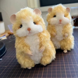 以下が含まれることがあります： 薄い茶色の毛並み、白いお腹、黒い目をしたぬいぐるみハムスターが2体。ハムスターは茶色の格子状の表面に立っています。