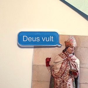 Könnte beinhalten: Eine blaue Sprechblase mit dem Text "Deus vult" befindet sich vor einer Statue einer religiösen Figur. Die Statue trägt ein detailliertes Gewand mit roten und goldenen Akzenten und einen verzierten Hut.