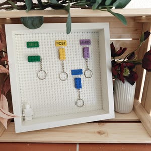 Könnte beinhalten: Eine weiße, gerahmte, Lego-kompatible Anzeigetafel mit Schlüsselanhängern, die an Lego-Steinen befestigt sind. Die Steine sind grün, gelb, blau und lila, jeder mit einem Namen. Die Anzeige befindet sich auf einem Holzregal mit einer Blumenvase.