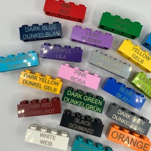 Può includere: Una collezione di blocchi di costruzione in plastica colorati con etichette in tedesco e inglese per ogni colore. I blocchi sono disposti a semicerchio con il blocco rosso in alto.
