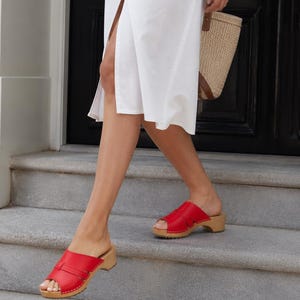 Puede incluir: Un par de zuecos de cuero rojo brillante con punta abierta, que presentan una tira ancha en el empeine, un detalle de hebilla pequeña y una suela de madera clara con tacón bajo. Los zuecos se llevan con un vestido blanco y un bolso de paja.