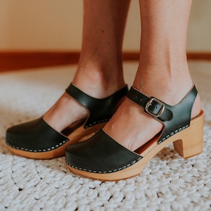 May include: Black leather clogs with a wooden sole and a block heel. The clogs have a strap across the top of the foot and an ankle strap with a silver buckle. The wooden sole is light brown, and the leather is stitched to the sole with white thread.