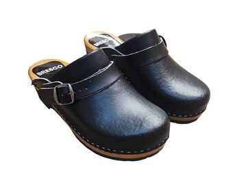 Puede incluir: Un par de zuecos de cuero negro con suela de madera y una correa con hebilla. Los zuecos son de la marca "BRESCO".