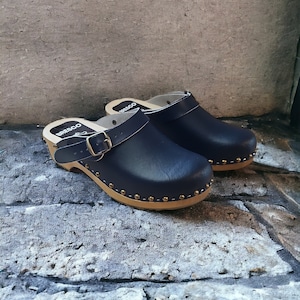 May include: A pair of dark blue leather clogs with a wooden base and a buckle strap. The clogs have a row of gold studs around the edge of the sole.