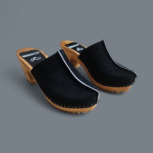 May include: Pair of black suede clogs with wooden heels and white stitching. The clogs have a white stripe down the center of the front and the brand name "BRESCO" is printed on the side.