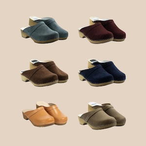 May include: Assortment of clogs in various colors, including denim blue, burgundy, brown, navy, tan, and taupe. Each pair features a wooden sole with a rounded toe and a suede or leather upper. The clogs are displayed on a neutral background.