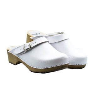 Könnte beinhalten: Weiße Ledersandalen mit Schnallenriemen und Holzabsatz. Die Sandalen haben einen genähten Rand und eine braune Holzsohle.